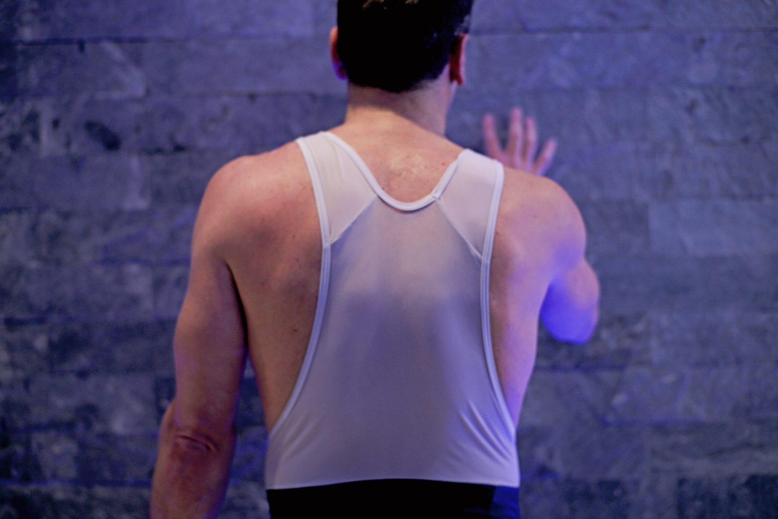 A man in a white tank top faces a dark stone wall, extending his right arm to touch it. His back muscles are visible, and the lighting casts a blue hue on the scene.