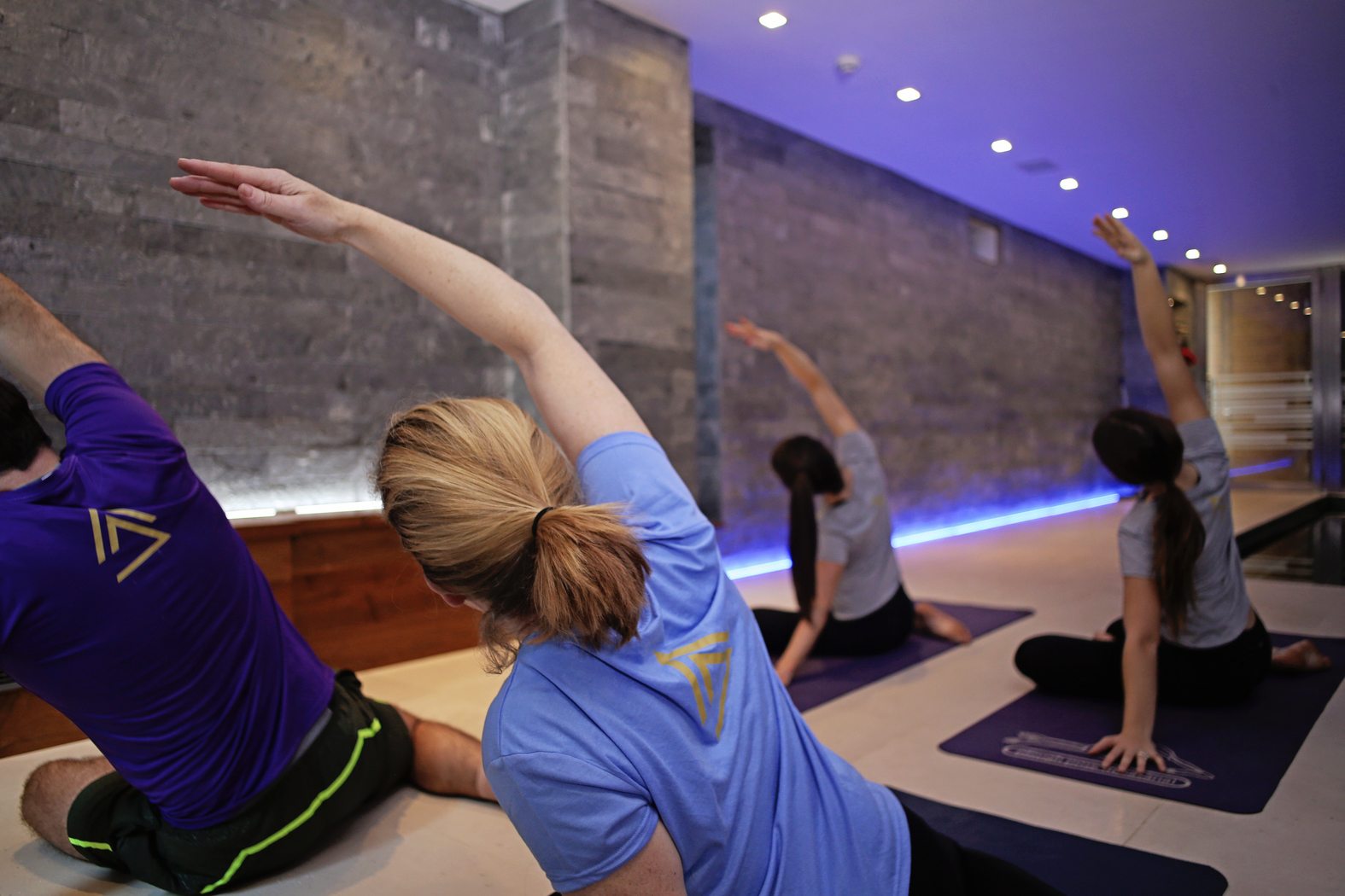 People in a yoga class performing side stretches on mats in a dimly lit room with concrete walls. The participants wear athletic clothing, and the lighting features a blue hue along the base of the wall.