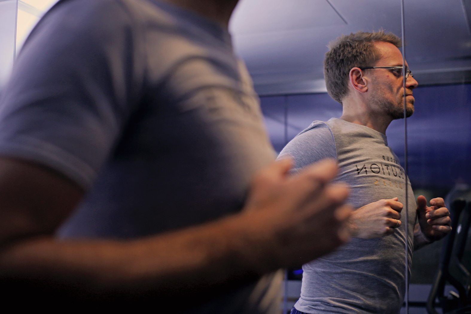 A person in a gray T-shirt is reflected in a mirror while running on a treadmill indoors. The focus is on their determined expression and posture, with a slightly blurred foreground of their torso.