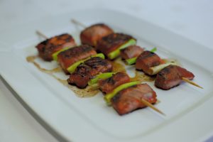 In the Cooking Demonstration, skewers of grilled meat paired with green vegetables are elegantly displayed on a rectangular white plate, enhanced by a delicious sauce and slightly charred to perfection for a savory look.
