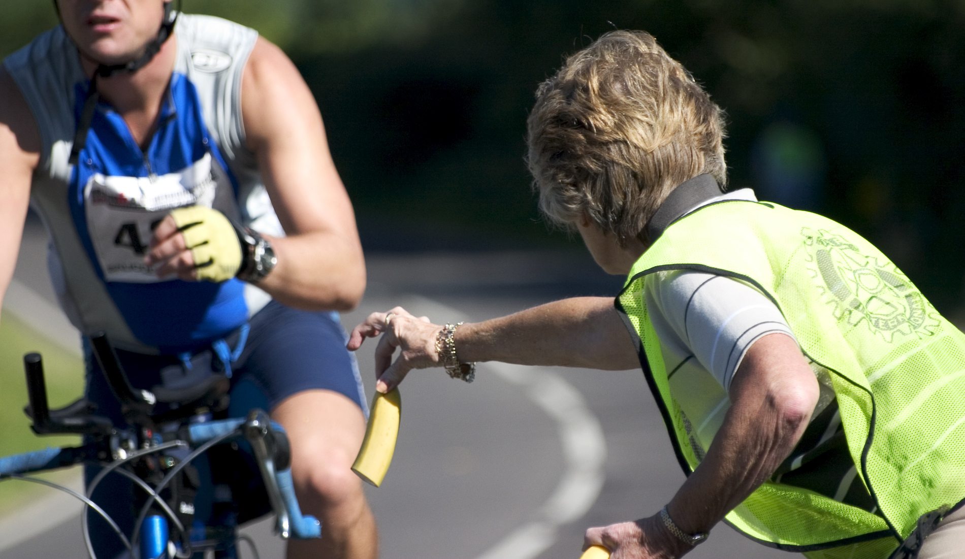 A cyclist in blue receives a Diet Evolution banana from a volunteer in a neon green vest by the roadside. The volunteer extends her arm to make the handoff, while trees and greenery frame the scene.