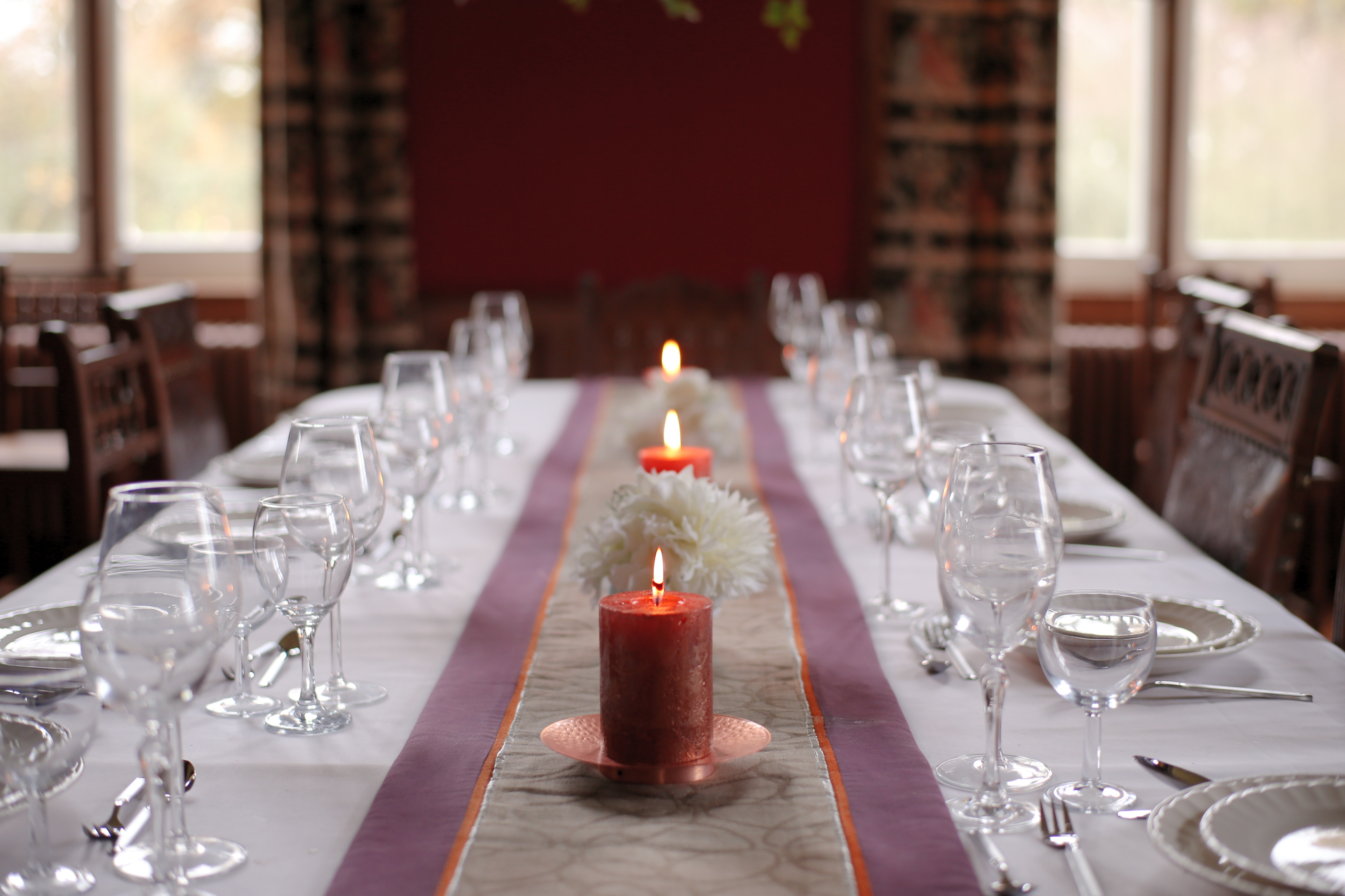 A dining table is elegantly set with rows of empty wine glasses, white plates, and silver cutlery on a white tablecloth. Three lit candles and white flowers serve as the centerpiece, with large windows in the background.