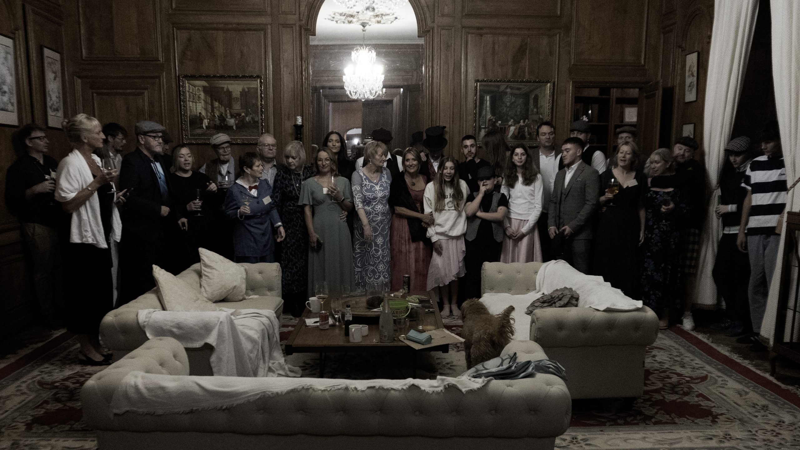 Dressed in vintage or formal attire, guests gather in an elegant wood-paneled room under a chandelier, surrounded by paintings. Some hold drinks near two cream sofas and a coffee table with glasses and a teapot. This scene is part of Death at the Chateau - Murder Mystery.