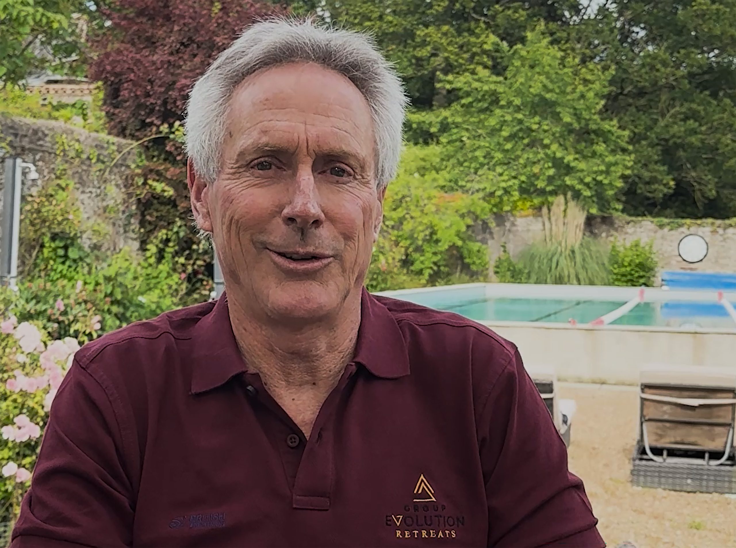 Mark Allen, wearing a maroon polo from the Mark Allen Experience training camp sits by the pool, with green foliage and a stone wall in the background.
